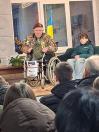 зображено українського волонтера дядька Гришу (Григорія Янченка) в інвалідному візку, який сидить у приміщенні з групою інших людей, зокрема жінками у зимовому одязі та чоловіком у пальті, тримаючий невеликий синьо-жовтий прапор