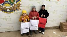 Троє дітей у зимовому одязі позують перед банером із символікою громади, тримаючи пакети з написом «BUFFETT».