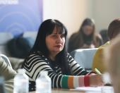 Жінка середнього віку з довгим чорним волоссям і смужчастим светром (чорно-білі смужки з кольоровими манжетами) сидить за столом, тримаючи ручку та дивлячись убік. На передньому плані видно пляшки з водою, а на задньому плані — інших людей і розмитий фон із синім банером