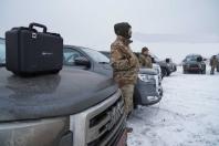 На зображенні великий чорний захисний кейс стоїть на капоті сірого пікапа на засніженій стоянці. Поруч стоїть чоловік у військовій формі та капюшоні, а на задньому плані видно інші автомобілі та людей