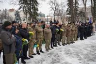 Урочиста церемонія або пам'ятний захід на відкритому повітрі взимку, де група людей, переважно чоловіків у військовій формі, стоять перед засніженим парком. Деякі з них тримають букети жовтих та синіх квітів, які символізують кольори українського прапора. На задньому плані видно будівлі та розмиті прапори.
