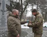 Чоловіки на зображенні беруть участь в урочистій церемонії на відкритому повітрі взимку. Один із них, чоловік у військовій формі (праворуч), тримає в руках нагрудний знак, який йому вручає чоловік у куртці кольору хакі (ліворуч). На нашивці на рукаві військового чітко видно літери «СБУ» (Служба безпеки України). На кам'яній стелі на задньому плані, біля якої відбувається подія, є написи, що включають дати «22.01.1918» та «22.01.2017», що, ймовірно, пов'язано з пам'ятною датою, можливо Днем Соборності Україн