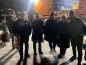 На зображенні група з п'яти людей у зимовому одязі стоїть на засніженій землі вночі перед яскраво освітленим помаранчевим фудтраком або великим автомобілем. Люди дивляться в камеру, а на задньому плані видно додаткове обладнання та ще одну особу ліворуч