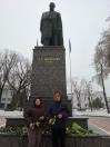 Двоє людей стоять біля пам'ятника Тарасу Шевченку взимку, тримаючи синьо-жовті квіти та стрічки. На задньому плані видно великий бронзовий пам'ятник на гранітному постаменті з написами: «Т. Г. ШЕВЧЕНКО» та датами «1814-1861». Біля основи пам'ятника лежать букети квітів. Навколо — засніжений парк та будівлі. Поруч майорить український прапор.