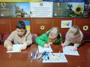 Троє дітей сидять за столом і малюють кольоровими олівцями та фломастерами. На задньому плані розташовані банери та плакати, зокрема один із написом «Координаційний центр надання допомоги внутрішньо переміщеним особам з Луганщини в Сумській області» та військові плакати із закликами приєднуватися до ЗСУ. Діти розфарбовують малюнки, а на стіні висить малюнок руки з українським прапором