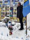 Чоловік у чорному одязі стоїть спиною до камери в засніженому місці, дивлячись на великий меморіальний стенд із фотографіями військових. На снігу видно невеликі українські прапорці та букет рожевих квітів.