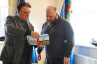 Зображення є фотографією двох чоловіків, які стоять у приміщенні з українськими прапорами на задньому плані. Чоловік ліворуч у картатому піджаку тримає відкриту книгу з яскраво-червоним корінцем та світлими сторінками, показуючи її чоловіку праворуч. Чоловік праворуч, із бородою, одягнений у сірий світшот, уважно дивиться на книгу.