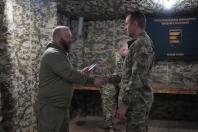На зображенні двоє чоловіків у військовій формі потискають один одному руки в приміщенні з камуфляжними стінами. У центрі сцени стоїть третій військовий, а на задньому плані видно синю табличку з текстом українською мовою: «ТРЕТЯ ПРИКОРДОННА КОМЕНДАТУРА ШВИДКОГО РЕАГУВАННЯ СРІБЛА ТРІЙКА»