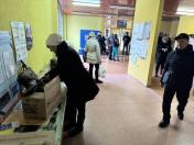 Зображення показує внутрішню сцену, схожу на центр видачі гуманітарної допомоги. На передньому плані ліворуч людина в чорному пальті та білій шапці схилилася над картонною коробкою з логотипом «GEM» (Global Empowerment Mission). Праворуч іде чоловік у чорній куртці та кепці. На задньому плані в коридорі видно інших людей та інформаційні плакати на стінах. Приміщення має яскраві жовті стіни та плиткову підлогу.