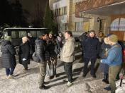 На зображенні група з приблизно десяти людей у різному зимовому одязі стоїть на засніженій землі перед входом до будівлі вночі. На задньому плані ліворуч також припарковано великий темний фургон