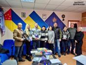На зображенні група з восьми людей позує в приміщенні з кількома прапорами, включаючи український, на стіні. Люди, одягнені в офіційний та повсякденний одяг, тримають книги, а на передньому плані розташований стіл із додатковими матеріалами. На стіні видно напис «ЗАХИСТ ДЕРЖАВИ"