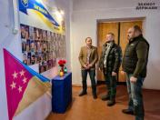 На зображенні троє чоловіків у приміщенні біля меморіальної стіни з багатьма портретами загиблих воїнів. На стіні також видно український прапор із написом «ЗАГИНУЛИ» та стрічку в синьо-жовтих кольорах, а в правому верхньому куті розташовано логотип «ЗАХИСТ ДЕРЖАВИ». На передньому плані ліворуч стоїть прапор із символікою, що нагадує прапор Щастинського району. Чоловіки одягнені в зимовий повсякденний одяг, включаючи дуті жилети та куртки.