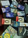 На зображенні велика колекція різноманітних книг розкладена на темній поверхні, схожій на стіл. Обкладинки книг мають різні кольори та дизайни. Серед помітних назв українською та англійською мовами є «ЖІНКА ВІЙНИ», «ЗОЛОТО НАЦІЇ», «WOMAN OF WAR» та «БАТАЛЬЙОНУ «ДОНБАС»». У правому верхньому куті також видно логотип «ЗАХИСТ ДЕРЖАВИ»