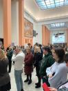 Зображення показує групу людей, які стоять у великому виставковому залі музею з колонами, фрескою та моделлю літака, що висить на стелі.
