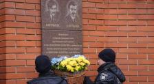 На зображенні двоє людей у формі української поліції стоять біля меморіальної дошки, встановленої на цегляній стіні, і тримають великий плетений кошик із жовто-блакитними квітами, щоб покласти їх на вшанування пам'яті діячів УНР братів Шульгиних