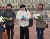 На зображенні три жінки стоять на брукованій площі перед великою історичною будівлею, одягнені в зимовий одяг і тримають квіти та синю свічку, беручи участь у церемонії вшанування пам'яті