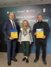 Три людини, двоє чоловіків у костюмах та жінка у вишиванці, стоять перед фоном із логотипами, тримаючи жовто-блакитні плакетки