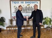 Двоє чоловіків тиснуть руки в кімнаті з великим плакатом на стіні з написом «Герої не вмирають...». 