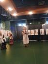 Жінка у вишитій сукні стоїть перед низкою виставкових стендів із зображеннями традиційного одягу та двома манекенами у фойє чи залі