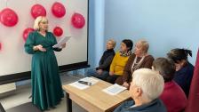 Ведуча у довгій зеленій сукні виступає з промовою перед групою жінок старшого віку в аудиторії. На фоні — білий екран, прикрашений червоними повітряними кульками
