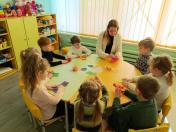 Учителька та група з семи дітей сидять за круглим столом у світлій ігровій кімнаті. Вони займаються творчою роботою, складаючи різнокольоровий папір. На задньому плані видно полиці з іграшками та велике вікно з жовтими жалюзі.
