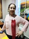 Усміхнена жінка в рожевому джемпері демонструє прикріплену до грудей червону прикрасу у формі серця з довгою бахромою, виконану в техніці макраме. Вона стоїть у приміщенні, поклавши руку на стегно.