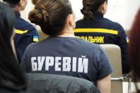 Жінка у футболці з написом «БУРЕВІЙ» сидить поруч із рятувальниками ДСНС під час офіційного заходу.