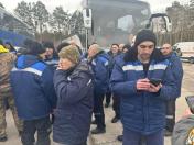 Група чоловіків у синьому робочому одязі, деякі з яких палять або користуються телефонами, стоять біля великого білого автобуса та автомобіля швидкої допомоги