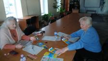 Три жінки сидять за столом у світлій бібліотеці, займаючись заповненням документів або творчою роботою з використанням кольорових олівців