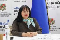 Жінка з блакитно-жовтою шийною хусткою сидить за столом з іменною табличкою «Катерина Безгинська» на фоні символіки Луганської обласної військової адміністрації.
