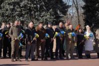 Група людей у цивільному та військовому одязі стоїть у шерензі під час урочистого заходу, тримаючи в руках букети квітів у синьо-жовтих кольорах державного прапора України.