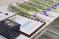 На білому столі підготовлені до вручення державна почесна грамота в рамці, стос посвідчень до нагород та ряд біло-фіолетових тюльпанів, перев'язаних стрічками