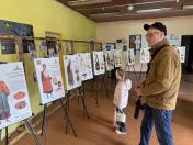 Чоловік у коричневій куртці та маленька дівчинка з білою сумочкою розглядають експонати виставки, присвяченої традиційному українському вбранню, де на мольбертах представлені інформаційні стенди з ілюстраціями та описами автентичних строїв.