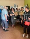 Група людей перебуває у приміщенні під час отримання гуманітарної допомоги, де жінка в довгій сукні та чоловік у кепці несуть картонні коробки з логотипами благодійних організацій, зокрема GEM, повз інших присутніх, що чекають у черзі