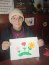 Усміхнена жінка в окулярах та світлій в'язаній шапці демонструє свій об'ємний малюнок із трьома різнокольоровими квітками, сидючи за столом у приміщенні координаційного центру допомоги переселенцям з Луганщини.
