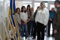 На світлині зображена група людей у світлому приміщенні біля скляних дверей із написом «Вихід», де на передньому плані стоять кілька дерев’яних мольбертів із картинами. Ліворуч видно державний прапор України та синій прапор Євросоюзу, а учасники заходу, серед яких чоловік у військовій формі та жінки в повсякденному одязі, зосереджено спостерігають за подіє