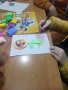 На світлині зображено процес дитячої творчості: дитина в жовтій куртці створює об'ємну аплікацію на аркуші паперу, використовуючи яскравий пластилін та олівці. Поруч на столі розкладено кольорові матеріали для ліплення та навчальні картки, а в центрі роботи — велика різнокольорова квітка.