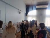Група жінок у світлому просторому залі уважно ознайомлюється з експозицією плакатів на триногах, які презентують історію та естетику традиційного українського строю через фотографії та детальні етнографічні описи.