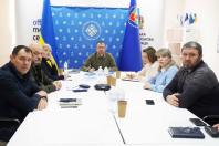 Навколо великого білого Т-подібного столу сидять семеро людей (чотири чоловіки та три жінки). Вони виглядають зосередженими, дехто дивиться прямо в камеру, інші — на документи чи спікера. У центрі столу, на фоні банера, сидить чоловік у військовій формі (кольору хакі).