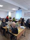 На передньому плані жінка в окулярах та світлій шапці уважно вивчає документ, поклавши поруч велику чорну сумку. Інші учасники також схилилися над столами, тримаючи в руках ручки.