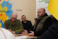 На світлині зафіксовано робочу зустріч трьох чоловіків за столом у приміщенні гуманітарного або адміністративного хабу, про що свідчить банер «Луганщина моя» на задньому плані. Один із присутніх у жилетці активно щось пояснює, жестикулюючи руками, тоді як інші зосереджено слухають та роблять нотатки