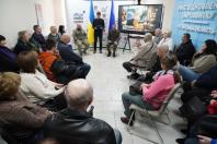 Фото зняте з ракурсу ззаду глядачів, які сидять півколом. У центрі кімнати стоять доповідачі, один з яких тримає папку. На стіні праворуч розміщено об’ємний напис блакитними літерами про офіс Луганщини. Велика увага прикута до монітора з яскравим колажем на тему Чорнобиля. Атмосфера офіційна та зосереджена.