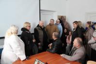 Робоча зустріч групи людей у невеликому приміщенні. Учасники обговорення стоять та сидять навколо дерев'яного столу, зосереджено слухаючи один одного. На задньому плані — великий екран для презентацій.