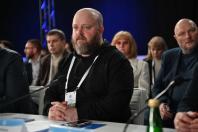 У центрі офіційного заходу за білим робочим столом сидить зосереджений бородатий чоловік у чорному худі з бейджем учасника на білій стрічці, оточений іншими делегатами в діловому вбранні на фоні конференц-залу з професійним синім освітленням.