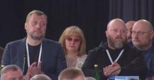 На передньому плані офіційного заходу зображено зосереджених учасників із бейджами на білих стрічках, серед яких виділяється чоловік у темно-синьому піджаку, що аплодує, та бородатий чоловік у чорному худі, які уважно спостерігають за подією на темному фоні зали.