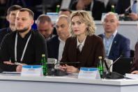 У центрі офіційного заходу за довгим білим столом із мікрофонами та табличками сидить зосереджена жінка у темно-коричневому костюмі з білявим каре поруч із чоловіком у чорному поло, оточені іншими учасниками у діловому вбранні на розмитому фоні конференц-залу.