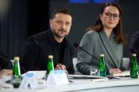 Під час офіційного засідання за білим столом із мікрофонами та скляними пляшками води зображено зосередженого Володимира Зеленського в темній куртці поруч із Юлією Свириденко в сірому діловому піджаку та окулярах, які уважно спостерігають за ходом зустрічі.