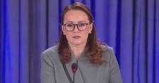 На тлі синіх вертикальних штор зображено зосереджену жінку в окулярах та сірому діловому піджаку, яка виступає перед мікрофоном під час офіційного заходу.