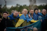 Група щиро усміхнених українських захисників, звільнених із російського полону під час великого великоднього обміну 11 квітня 2026 року, позує для фото, загорнувшись у національні прапори та тримаючи стяг із символікою морської піхоти та девізом «Вірний завжди».