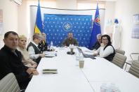 У світлому офісному приміщенні група посадовців проводить робочу нараду за довгим білим столом, на чолі якого сидить чоловік у військовій формі на фоні синього банера та прапорів України й Луганської області.